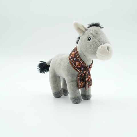 A small plush donkey toy standing on a seamless white studio background. The donkey is light gray with a cream muzzle and ears, dark gray hooves and tail, and a patterned brown and orange scarf around its neck.