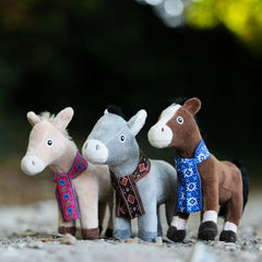 Three small plush toys standing outdoors on a gravel path with a blurred green and dark background. There is a mule, donkey and horse all wearing scarves.