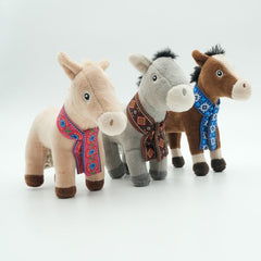 Three small plush toys standing side by side on a clean white studio background. There is a mule, donkey and horse, each wearing a colourful embroidered scarf.